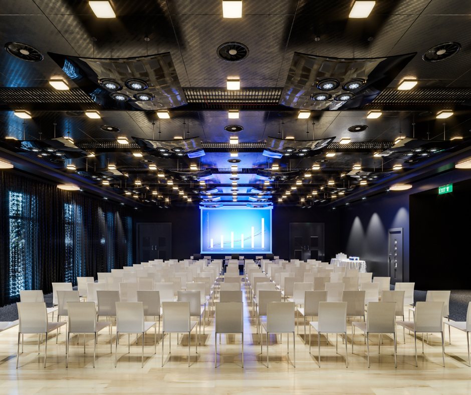 Image of a conference room shot from behind, showing rows of white chairs and a stage in the centre.